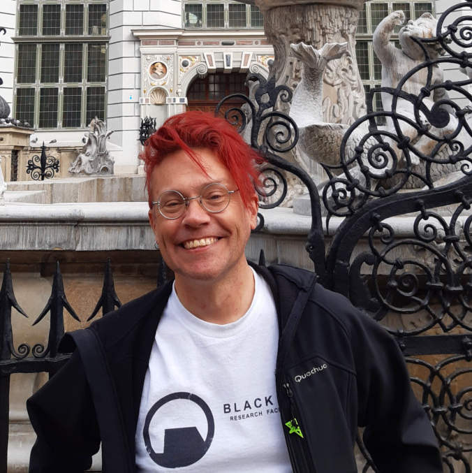 Niall (a white man with dyed red hair) stood in front of an ornate carved fountain.